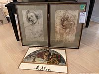 Two framed prints of classical-style drawings and a hanging art book with classical figures artwork and signature on floor