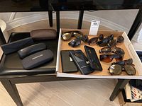 Box with nine pairs of sunglasses and three phone cases inside, and four glasses cases outside the box on chair