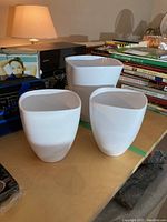 Three white ceramic vases of varying sizes placed on a table among books and other household items.
