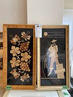 Photo of two wood laminate art pieces framed behind glass in wood frames, showing floral grapes design and a figure fishing under moonlight.