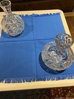 View of both crystal cut decanters on blue cloth on a table, showing their overall shape and size difference