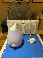 Photo showing the Crane humidifier, Lasko window fan, and power adapter on a blue cloth.
