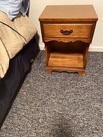 Front view showing the drawer with metal handle and open shelf below, positioned beside a bed on carpet flooring.