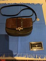 Brighton One World crossbody bag and Love Beat card holder on blue fringed cloth, showing bag's front with brown crocodile pattern flap and silver heart clasp and separate card holder with silver heart emblem.