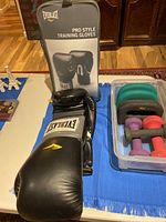 View of black Everlast Pro Style training gloves displayed next to a clear plastic container holding various colorful hand weights on a blue surface.