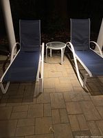 Two navy blue chaise lounges with white metal frames and a white round table with textured glass top between them on stone patio.