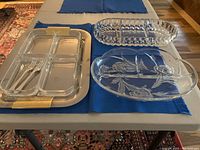Wide view of all three relish dishes placed on a blue table runner, showing the stainless steel dish with glass inserts and two forks on the left and two cut glass dishes on the right.