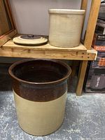 Two ceramic crocks placed near a wooden shelf, one with lid removed and visible interior, cream and brown coloring.