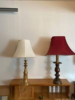 Both lamps side by side on wooden table against white wall showing the brass base lamp with off-white shade and wood base lamp with red shade.