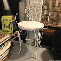 Angle front-left view of vintage metal framed vanity chair with round white cushioned seat showing structural frame, armrests and backrest details.