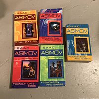 Photo shows five paperback Isaac Asimov Foundation series novels laid out on a gray concrete floor. Titles visible are Foundation, Forward the Foundation, Foundation's Edge, Foundation and Empire, and Foundation and Earth. Each book cover features distinct cover art and different background colors.