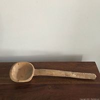 Full view showing the entire length of the oversized wooden spoon ladle on a wooden surface against a white background.