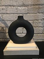 Black round vase with hollow circular center and narrow top opening, placed on a white surface against a brick wall background.