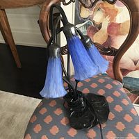 Full view of vintage glass lamp showing three blue flower bell-shaped glass shades and black metal leaf base, lamp placed on a patterned chair.