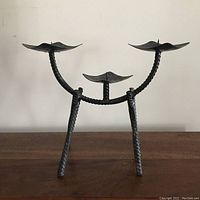 Front angled view of black iron candelabra showing three legs, curved arms, and leaf-shaped candle holders with patina and signs of wear.