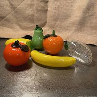 Five vintage Murano style glass fruit figurines arranged on a surface with a burlap background, showing variety of colors and shapes including oranges, pear, banana, and lemon.