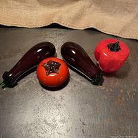 Four Murano glass style vegetable figurines including two eggplants, one tomato and one red bell pepper on a metal surface.