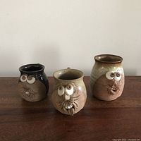 Three small decorative studio pottery mugs with sculpted owl or grotesque faces shown from the front. Each mug is different glaze color and style.