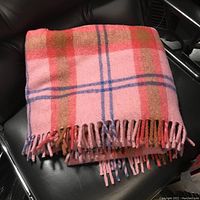 Folded pink wool blanket with plaid pattern placed on black leather chair, showing fringed edges.