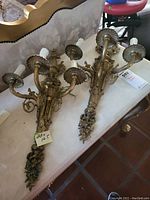 Pair of two brass wall sconces laid on a marble surface, showing both sconces with three arms and candle-style bulbs, ornate detailing visible