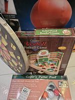 Photo showing boxed Golfer's Putter Pool game, Classic Baseball Pinball Game box, dartboard edge and orange exercise ball behind games.