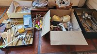 Overview of kitchen utensils, tools and gadgets arranged in trays and cardboard box, showing wooden spatulas, metal utensils, scissors, can opener, peelers and kitchen accessories.