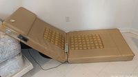 Beige padded massage bed laid on floor, showing angled fold and massage node area on the back section along with wired remote control