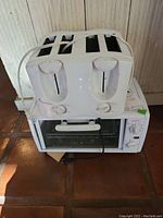 White four-slice Toastmaster toaster with two control knobs, sitting on top of a white Sunbeam toaster oven on tiled floor against wood paneling background.