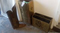 Photo showing a pair of brass umbrella boots shaped as an umbrella stand and a rectangular brass magazine holder with embossed designs placed on the ground next to a door.