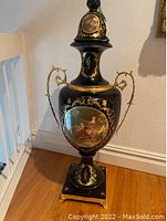 Black urn with gold decoration and painted classical scene on front. Scroll-like gold handles attached on sides. Square base with small feet. Visible damage on lid.