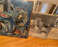 Three pieces of wall art including framed magazine print, screen printed building, and acrylic painting arranged leaning against wall on wood floor.