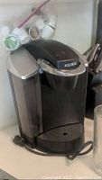 Front view of black and silver Keurig coffee maker with water reservoir and power cord visible.