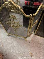Large brass/gold colored metal fireplace screen with decorative floral elements and mesh, with curved feet on floor