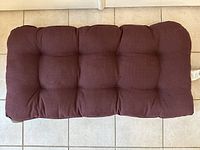 Front view of one burgundy tufted outdoor cushion for wicker settee on tiled floor.