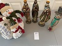Photo showing three Wise Men candlestick holders standing upright and two snowman decorations including a knit-style snowman and a glitter snowman.