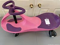 Overall front and side view of the Plasma Car on a table showing its pink body with purple seat and handles, featuring four small wheels underneath for smooth riding.