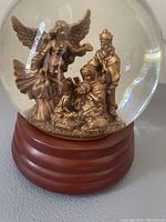 Front view showing the nativity scene figures inside the clear snow globe on a wooden base, including an angel, Mary, Joseph, baby Jesus, and a wise man.