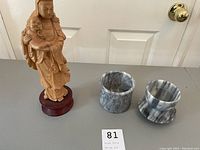 Photo showing carved wood statue with robed figure holding child on wooden base, alongside two pieces of gray and white marbled pot