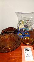 Four different sized amber glass ashtrays on table with plastic bag of assorted matchbooks and two blue Camel card decks visible