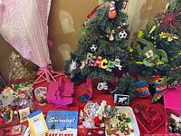 Wide view showing three tabletop artificial Christmas trees, assorted ornaments, a decorative plate, and other holiday decor items spread on a surface