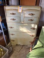 Full front view of the tall dresser showing seven drawers and gold-tone decorative handles and trim accents. The dresser is painted light green and has visible scratches.