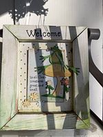 Wooden framed sign with a 'Welcome' message, featuring a 3D frog figure and flowers with text 'to all God's creatures great and small.'