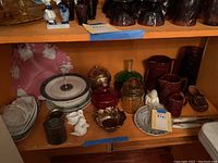 Shelf filled with assorted glass and ceramic serving ware including plates, jars, pitchers, and decorative items organized on one shelf.