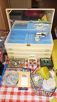 Photo showing large cream-colored jewelry box with blue velvet interior and mirror, surrounded by various decorative tins and cases on a red patterned cloth.