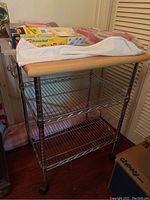 Front view of metal rolling kitchen cart with wood top and party supplies on the cart including napkins, plates, plastic wrap and doilies.