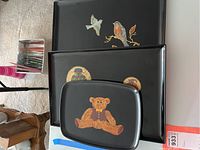 Three black lacquered platters stacked with decorative motifs of two birds, woven baskets, and a teddy bear.