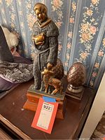 Full view of metal sculpture showing man holding animal and dog standing to side on wooden base.