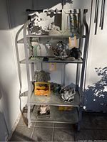 Outdoor metal shelving unit with four glass shelves shown with various decorative items on shelves. Unit has light gray metal frame and glass shelving with visible dirt and wear. Located against wall on tiled floor.