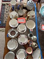 Overhead view of assorted teapots, cups, saucers, and pitchers on wooden table with various floral and insect designs, some stacked cups, and a mix of colors and patterns.