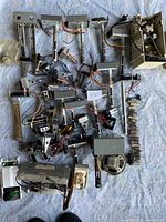 Photo showing a large assortment of vintage electrical pieces and parts laid out on a light blue fabric background. Items include multiple metal switches, attached wires, mounting brackets, and transformers.
